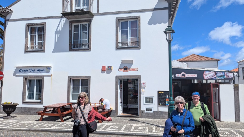 Un groupe de trois personnes devant un bâtiment blanc avec des fenêtres en bois. Un homme et une femme sourient, tandis qu'une autre femme prend une pose amusante. À l'arrière-plan, on voit une petite rue avec une lampe publique et une boutique.